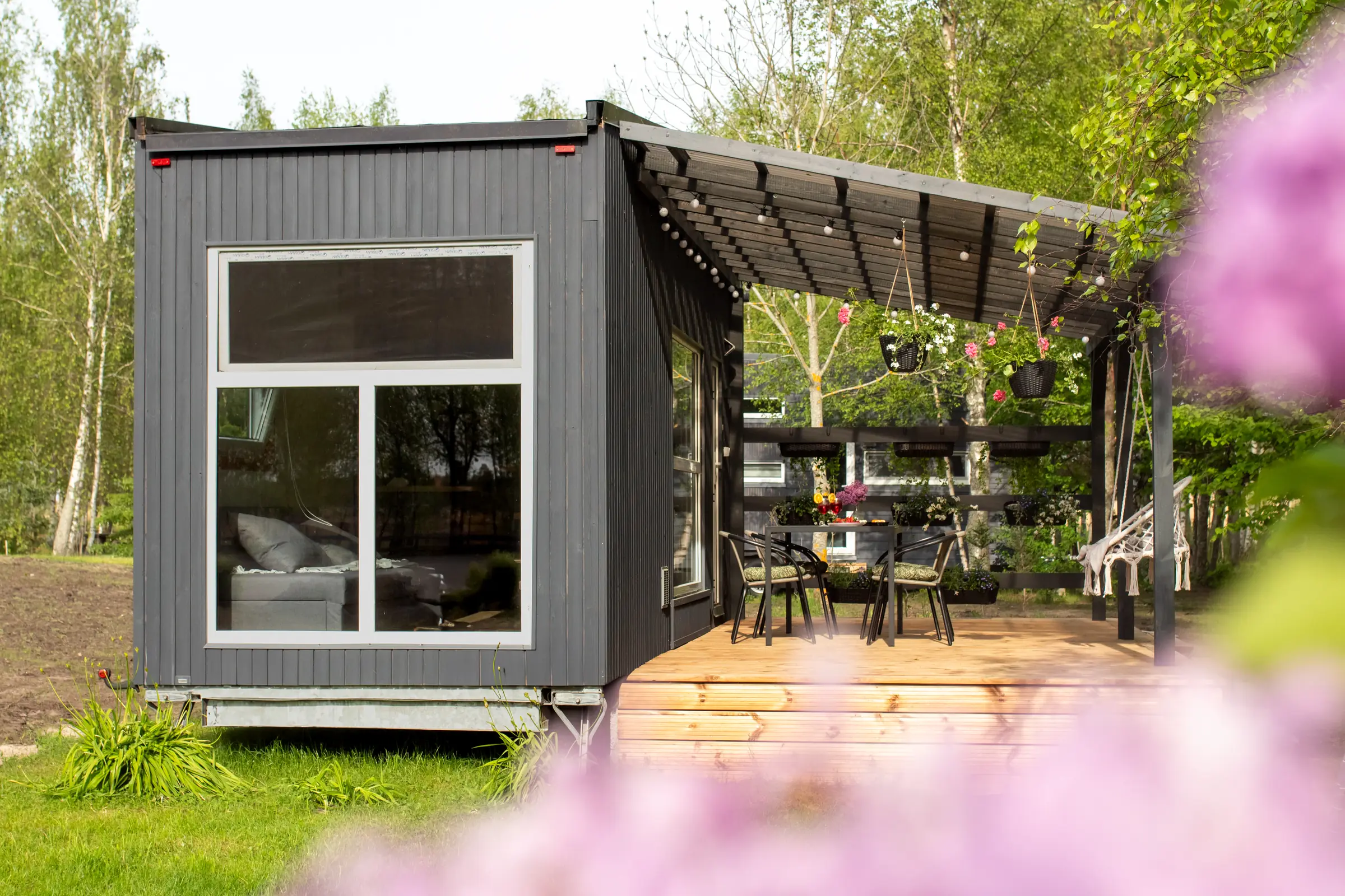 Saulriets no priekšpuses: liels logs, pergola ar lampiņām un terase